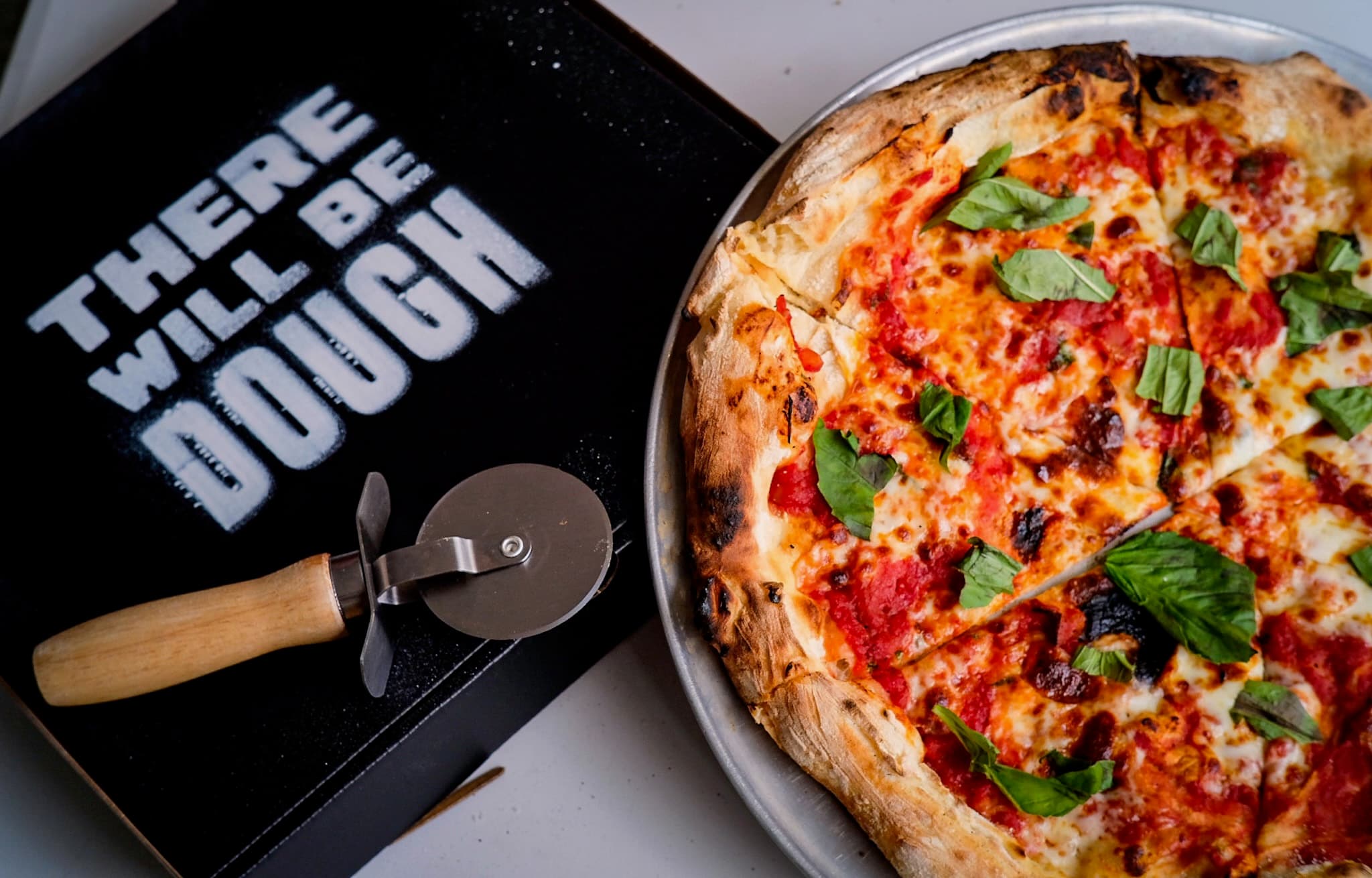 Margherita next to a There Will Be Dough box and a wooden pizza cutter, top-down